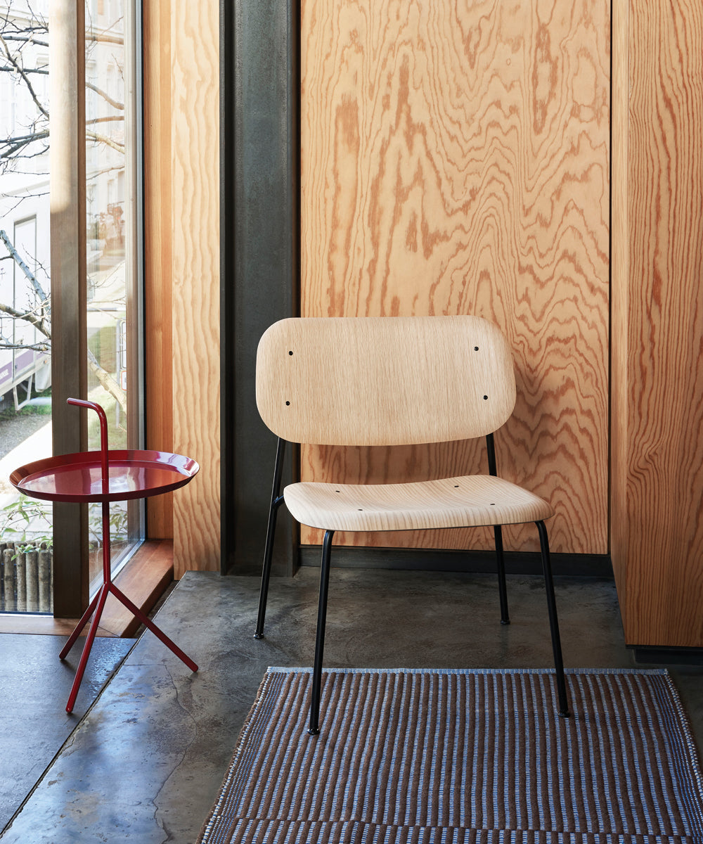 Light wood lounge chair with black metal legs beside a red side table, set against warm wood-paneled walls and a large window.